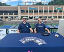 Two men sit at an outdoor table with brochures, in front of tennis and pickleball courts at a sports center.