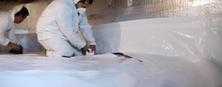 Two workers in protective suits install vapor barrier sheets in a crawl space.