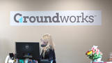 A person with a headset works at a computer under a "Groundworks" sign, next to a bouquet of colorful flowers.