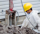 Worker in a yellow helmet and white suit operates drilling equipment near a building foundation.