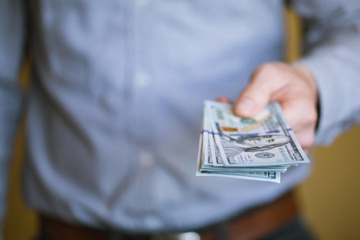 A person in a blue shirt holds out a stack of U.S. dollar bills, extended towards the camera.