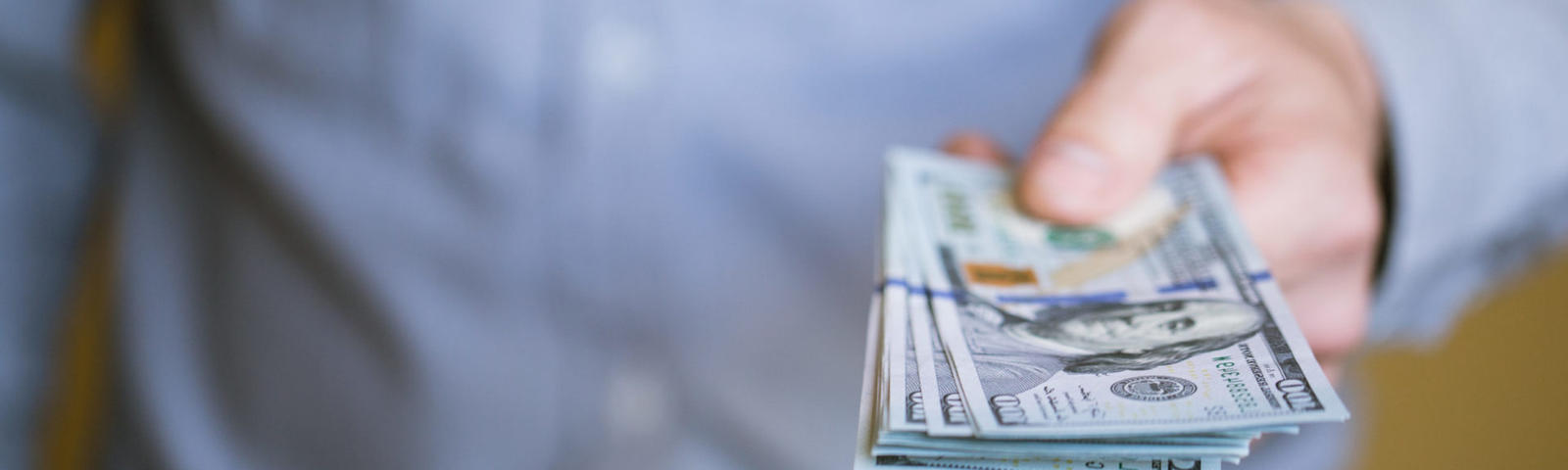 A person in a blue shirt holds out a stack of U.S. dollar bills, extended towards the camera.