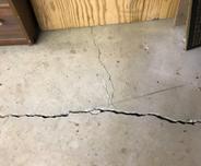 A large crack runs across cracked floor slabs near a wooden wall and furniture.
