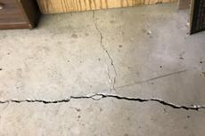 A large crack runs across cracked floor slabs near a wooden wall and furniture.