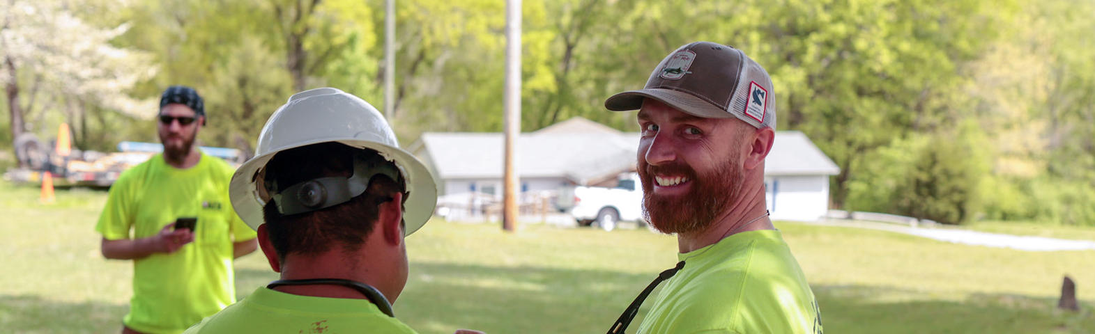 Three men outdoors wearing neon shirts and hats, two facing away and one smiling at the camera.