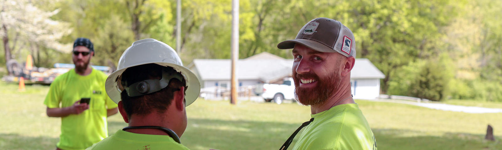Three men outdoors wearing neon shirts and hats, two facing away and one smiling at the camera.