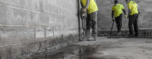 Foundation specialists in bright shirts expertly maneuver tools on a concrete floor inside an unfinished building, seamlessly working amid reflective water puddles.