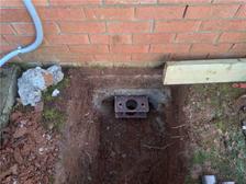 A dug hole near a brick wall with an underground metal bracket exposed and some debris around.