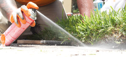Person in gloves sprays foam sealant and termiticides along the edge of grass near a sidewalk in bright sunlight.
