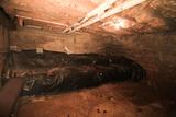 A dimly lit basement with exposed pipes, insulation, and a mound covered in plastic sheeting gives off typical crawl space smells.