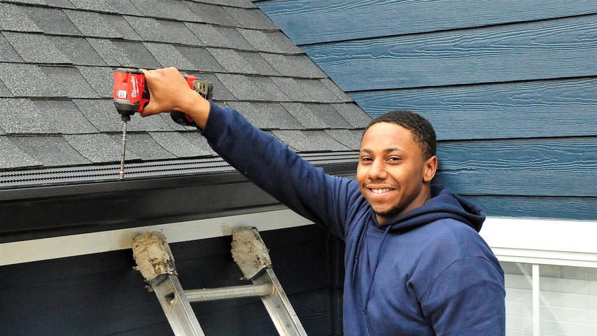 On a crisp Chattanooga morning, a person in a blue hoodie stands on a ladder, cheerfully using a drill to fix roof shingles.