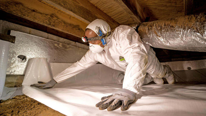 AFS crew member installing vapor barrier in crawlspace while wearing protective gear.