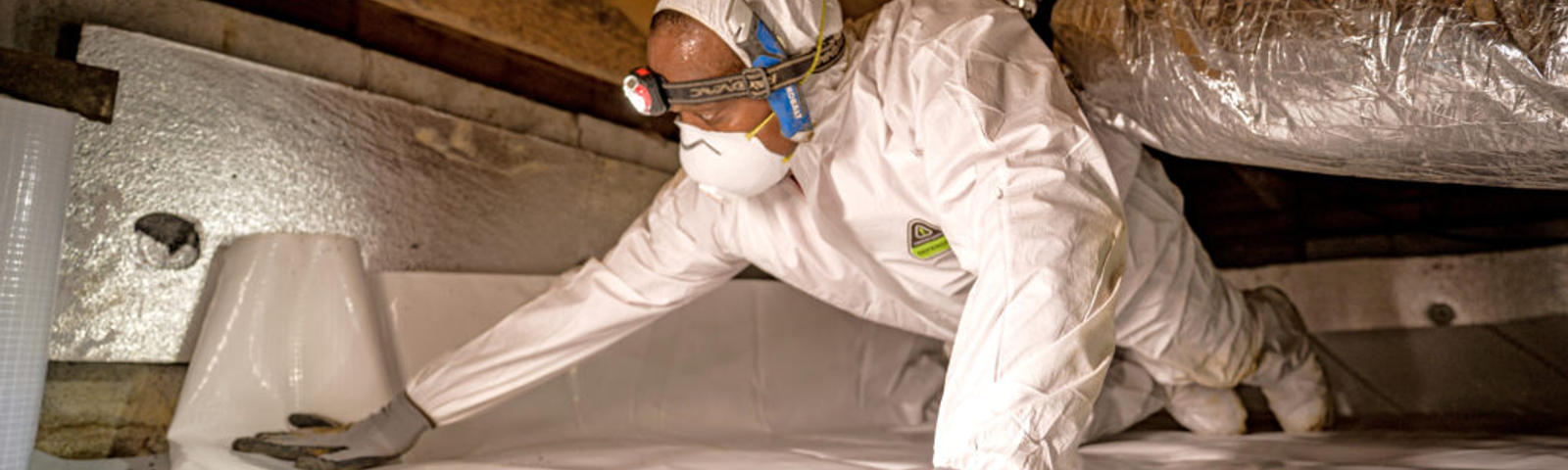 AFS crew member installing vapor barrier in crawlspace while wearing protective gear.