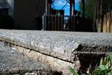 Close-up of cracked, uneven concrete sidewalk—perfect for an Alabama concrete repair project—with grass edging and a house in the background.