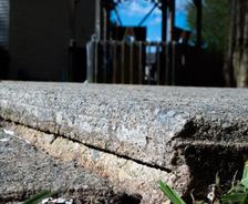 Close-up of cracked, uneven concrete sidewalk—perfect for an Alabama concrete repair project—with grass edging and a house in the background.