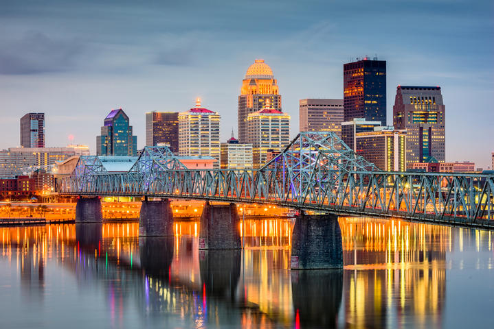Evening skyline of a city with tall buildings, a lit bridge, and reflections on a calm river.