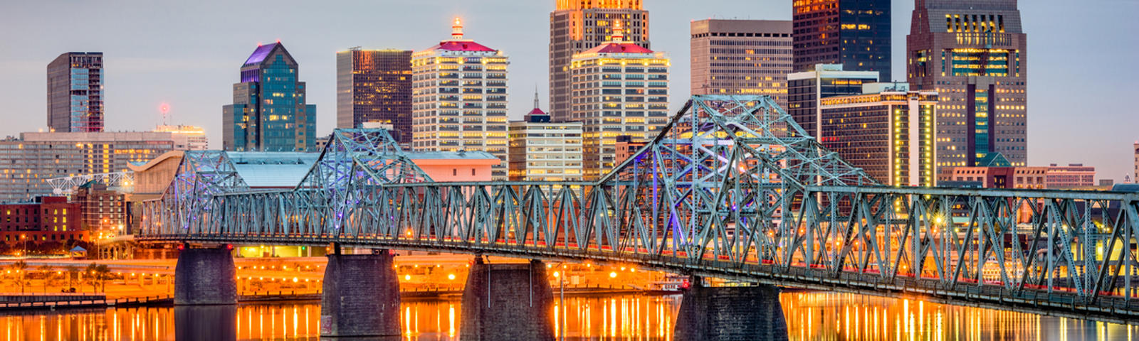 Evening skyline of a city with tall buildings, a lit bridge, and reflections on a calm river.