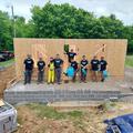 Group of people in matching shirts posing at a construction site with a wooden wall and greenery in the background.
