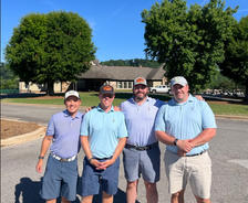Four men in golf attire standing on a sunny day with trees and a house in the background.