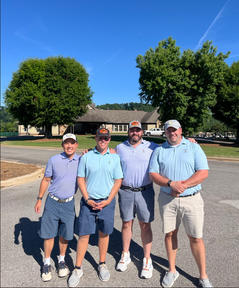 Four men in golf attire standing on a sunny day with trees and a house in the background.