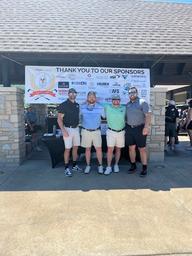 Four men pose in front of a sponsor banner at an outdoor event near a stone building.