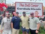 Four men pose in front of a banner for "The Bell Center Cornhole Classic" at an outdoor event.
