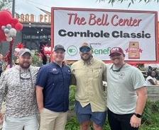 Four men pose in front of a banner for "The Bell Center Cornhole Classic" at an outdoor event.