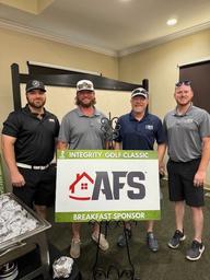 Four men in AFS outfits standing by a "Integrity Golf Classic" breakfast sponsor sign indoors.