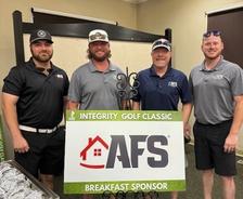 Four men in AFS outfits standing by a "Integrity Golf Classic" breakfast sponsor sign indoors.