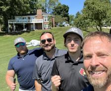 Four men smiling and posing with golf clubs on a sunny day at a golf course, with trees and a house in the background.