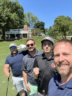 Four men smiling and posing with golf clubs on a sunny day at a golf course, with trees and a house in the background.