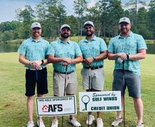 Four men in matching golf shirts stand with clubs by sponsor signs on a golf course.
