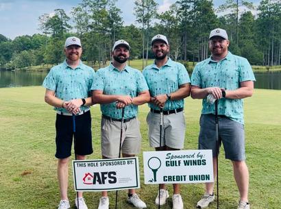 Four men in matching golf shirts stand with clubs by sponsor signs on a golf course.