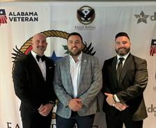 Three men in formal attire standing in front of banners for Alabama Veteran and Eagle Rising Veteran Consultants.