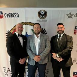 Three men in formal attire standing in front of banners for Alabama Veteran and Eagle Rising Veteran Consultants.