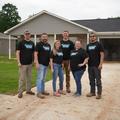 Six people wearing matching shirts stand in front of a single-story house on a driveway.