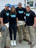 Four people wearing matching "Build Together" shirts pose in front of a construction site.