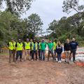 A group of people in casual and reflective clothing stand outdoors near a construction site surrounded by trees.