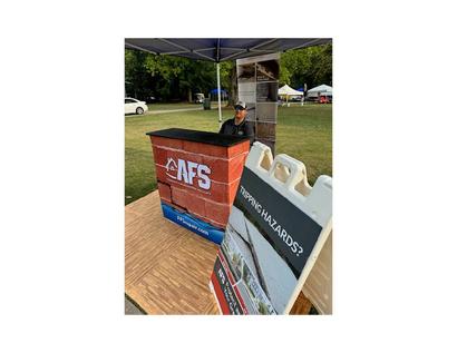 Man sitting at an AFS booth with a "Tripping Hazards?" sign at an outdoor event.