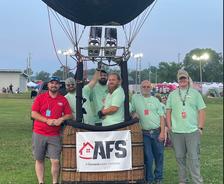 A group of five people stands beside a hot air balloon basket in a grassy area, soaking in the vibrant atmosphere of the Alabama Jubilee.