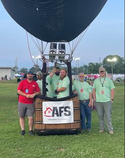 A group of five people stands beside a hot air balloon basket in a grassy area, soaking in the vibrant atmosphere of the Alabama Jubilee.