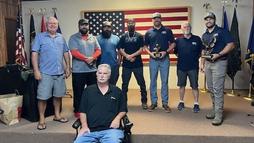 Seven men stand in front of a large American flag, holding trophies, with one man seated in front.
