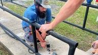 Man kneeling, working with a power tool on a concrete surface, while another person holds a railing.