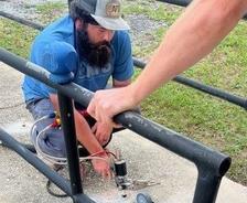 Man kneeling, working with a power tool on a concrete surface, while another person holds a railing.