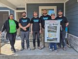 A group of six people stand on a porch holding a framed sign, wearing construction-related clothing and smiling.