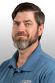 Man with a beard wearing a blue collared shirt against a gray background.