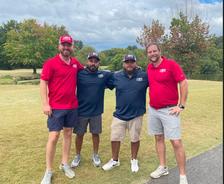 Four men in golf attire standing on a golf course with trees in the background.
