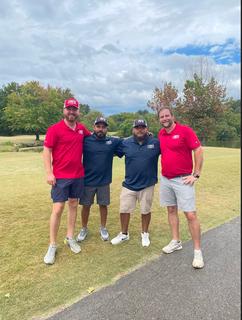 Four men in golf attire standing on a golf course with trees in the background.