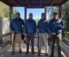 Four people in blue jackets holding rifles, standing on a wooden platform outdoors.