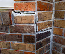 Cracked red and brown brick wall with visible mortar damage and repairs, located at a corner of a building.
